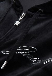 Hoodie preto em veludo com um puxador de zíper em forma de flor prateada e aplicações de strass num padrão ondulado ao longo de um dos lados. Textura macia.