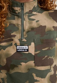 Grön kamouflagepullover med hög krage, dragkedja fram, en ficka med lappar och en Adidas-logotyp på bröstet.