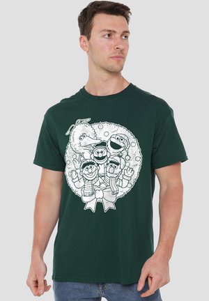 Mann in einem dunkelgrünen T-Shirt mit einem schwarz-weißen Weihnachtskranz, der fünf Figuren aus der Sesamstraße zeigt.