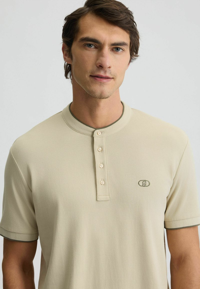 Tricou polo bej cu mânecă scurtă, realizat dintr-un material texturat, cu guler rotund și trei nasturi la decolteu, având accente verzi pe guler și pe mâneci.