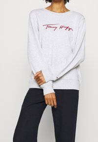 Světlě šedá mikina z bavlněné směsi. Má kulatý výstřih, dlouhé rukávy a červeně vyšitý nápis "Tommy Hilfiger" na přední straně.