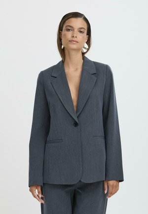 Femme portant un blazer gris foncé sur mesure et un pantalon assorti, avec de grandes boucles d'oreilles argentées, debout devant un fond clair uni.