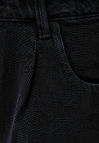 Svarta jeans av denim visar en närbild av tygets strukturerade yta, sömmar och fickdetalj. Materialet verkar robust och hållbart.