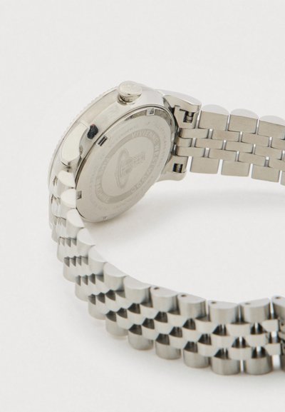 Sudraba krāsas metāla rokaspulkstenis ar saistītu aproci, uz balta fona redzama gravētā aizmugure un regulēšanas vainadziņš.