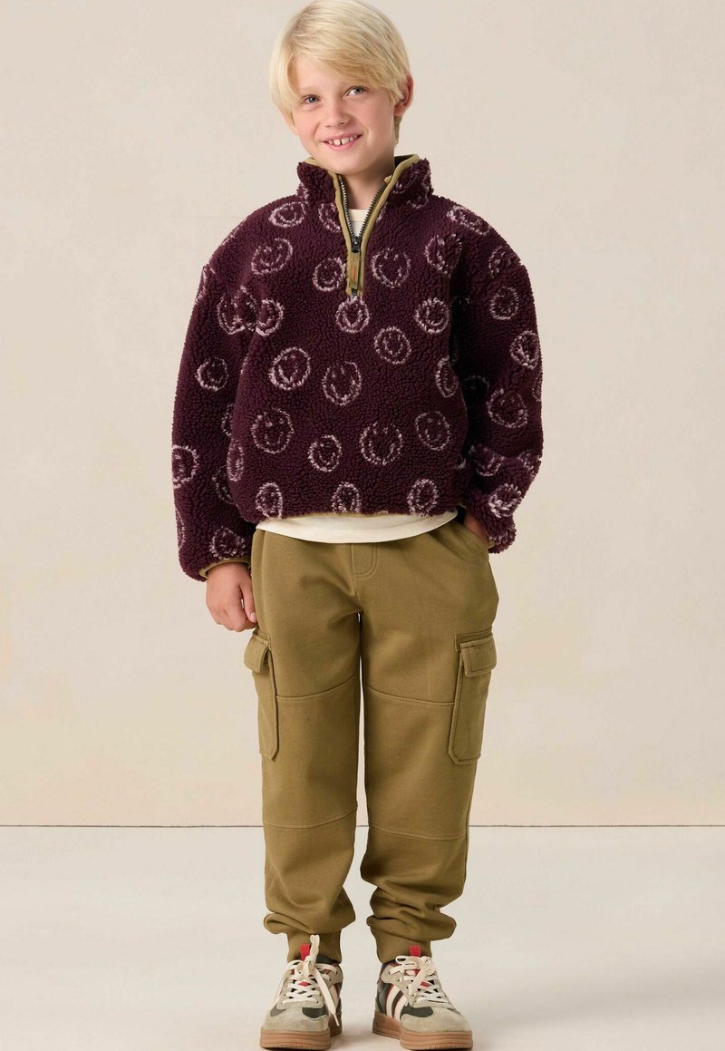 Paarse fleece pullover met smiley face patroon, kwart rits, gecombineerd met lichtbruine cargobroek met zijzakken en beige sneakers.