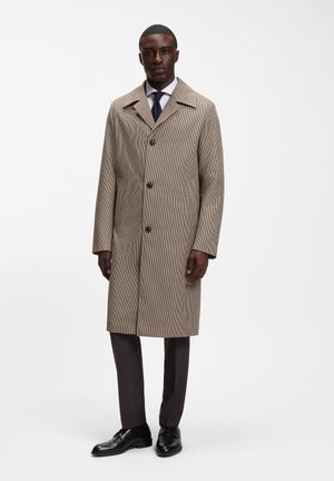 Mand iført en beige ternet frakke med tre knapper, hvid skjorte, mørkt slips, mørke bukser og sorte pæne sko på hvid baggrund.
