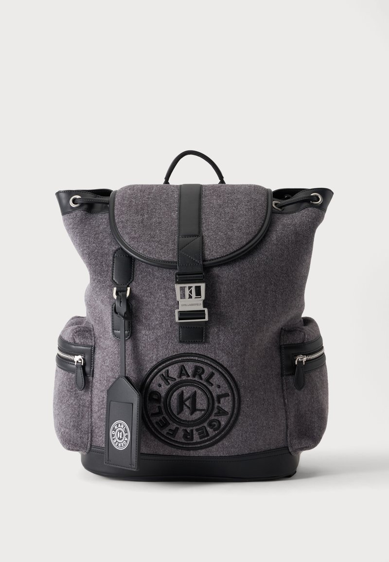 Sac à dos en feutre gris et noir avec rabat avant, deux poches latérales zippées, une poignée supérieure, une boucle à l'avant et un logo circulaire Karl Lagerfeld.