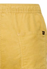 Gule manchesterbukser med elastisk linning og rektangulært baglomme med et lille sort og guld label.