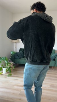 Persona che indossa una giacca nera e jeans azzurri chiari, in piedi in un soggiorno con un divano verde, piante d'appartamento e una lampada da terra.