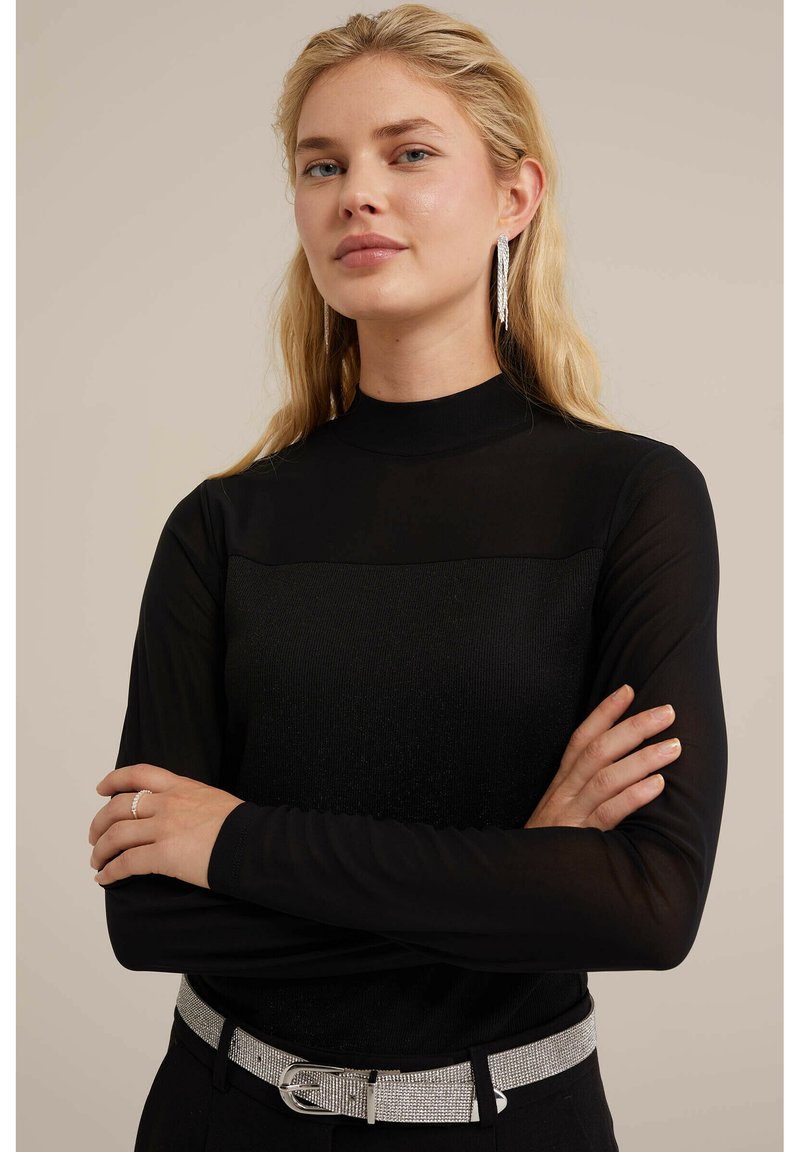 Zwarte lange mouwen top met een textuurpaneel en een hoge hals. Gekocht met een zilveren glitterriem. Model heeft licht golvend haar en oorbellen.
