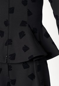 Černý peplum top z texturované látky, zdobený jemným vzorem černých tvarů papírových letadel. Zapínání na zip na přední straně.