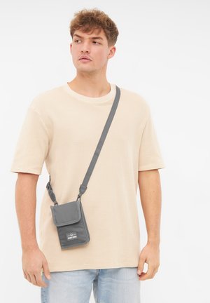 Szara torebka typu crossbody wykonana z teksturowanej tkaniny, z klapką i regulowanym paskiem, noszona na jasnobeżowej koszuli.