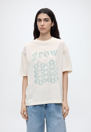 Frau mit langen dunklen Haaren, die ein übergroßes, elfenbeinfarbenes T-Shirt mit hellem blauen "grow"-Schriftzug und hellblaue Jeans trägt.
