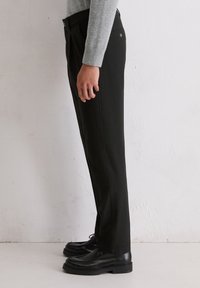 Pantalons noirs à coupe droite, fabriqués en tissu texturé, associés à des chaussures noires à lacets, présentés de côté sur un fond neutre.