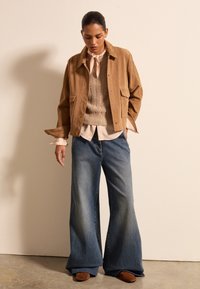 Tannfarbenes Wildlederjackett mit zwei Fronttaschen, kombiniert mit einem strukturierten beigen Pullover und weitem blauen Denim-Jeans. Braune Wildlederschuhe runden das Outfit ab.