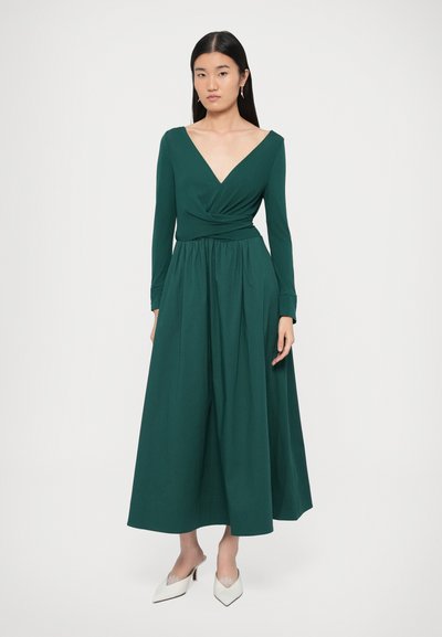 Φόρεμα με μακριά μανίκια, βαθύ V-λαιμόκοψη και τύλιγμα σε σκούρο πράσινο ύφασμα, με σφηκοφωλιά στη μέση και αέρινη φούστα. Συνδυασμένο με λευκά ψηλοτάκουνα παπούτσια με μυτερή μύτη.
