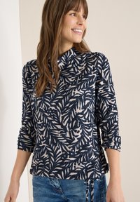 Haut à motifs bleu marine et crème avec un col montant, présentant des dessins de feuilles de palmier, des manches trois-quarts et un détail de nœud sur le côté.