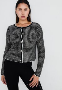 Svart och vit mönstrad cardigan med rund hals, långa ärmar och framknappar. Har kontrasterande vit kant längs kanterna.
