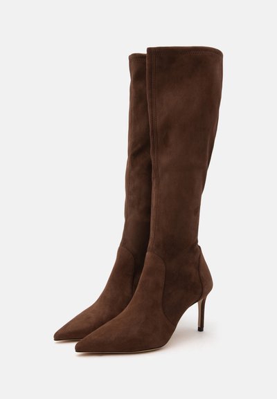 Paire de bottes hautes en daim marron à bout pointu avec des talons hauts fins, debout sur un fond blanc.