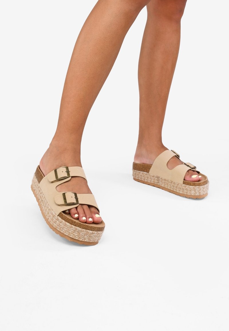 Jambes portant des sandales à plateforme beige avec deux brides réglables à boucle et des semelles tressées style espadrilles sur fond blanc.