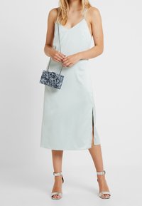 Abito in satin verde menta chiaro con spalline sottili e spacco laterale, abbinato a una pochette blu a fantasia e sandali argentati con cinturino alla caviglia.