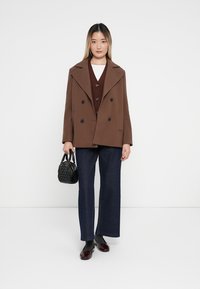 Brun dobbeltspent frakk med store reverser, over en bordeaux vest og hvit skjorte. Vide, mørke denimbukser, svart håndveske, patent sko.