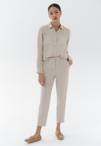 Chemise beige à boutons avec deux poches poitrine, assortie à un pantalon beige clair ajusté et des mocassins marron clair. Texture de tissu lisse.