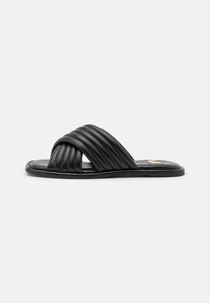 Sort slide-sandal med polstrede krydsede remme og flad sål på en ensfarvet hvid baggrund.