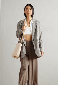 Prešívaný oversize blejzer v sivej a bielej farbe, kombinovaný s bielym crop topom a hnedými širokými nohavicami; drží béžovú tkanú kabelku.
