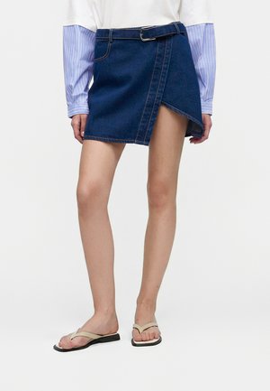 Person iført en mørkeblå denim omslagsnederdel med bælte, hvid skjorte med blåstribede ærmer og beige klip-klappere, står på en ensfarvet baggrund.