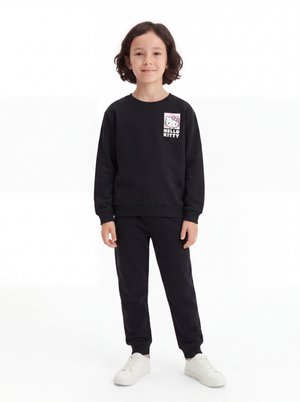 Barn med skulderlangt mørkt hår iført sort Hello Kitty-sweatshirt, sorte bukser og hvide sneakers, stående på hvid baggrund.