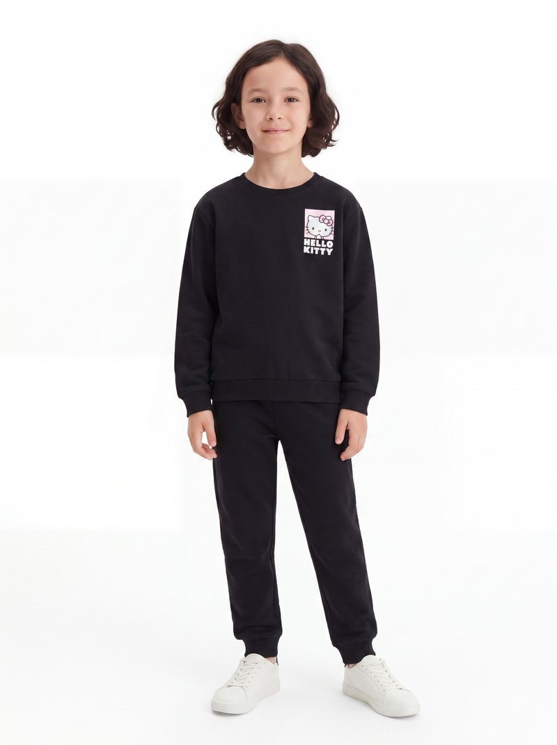 Enfant aux cheveux foncés mi-longs portant un sweat-shirt Hello Kitty noir, un pantalon noir et des baskets blanches, debout sur un fond blanc.