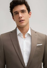 Brauner maßgeschneiderter Anzugjacke mit Revers, kombiniert mit einem weißen Hemd. Verfügt über eine Brusttasche mit einem weißen Taschentuch. Glatte Stoffoberfläche.