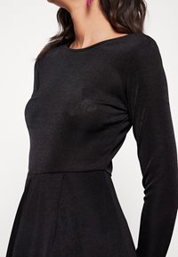 Robe noire à manches longues avec un corsage ajusté et une jupe évasée. Fabriquée dans un tissu lisse, elle présente une texture subtile et un col rond.