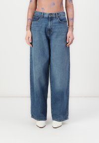 Lys blå denim brede bukser med høy midje, med fem lommer og knappelukking, kombinert med hvite spisse sko.