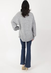 Übergroßer grauer Strickpullover mit geripptem Saum, kombiniert mit dunklen Schlagjeans. Glatte Textur, lockere Passform und lange Ärmel.