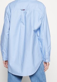 Light blue button-up shirt with a relaxed fit, back pleat detail, and rounded hem. Features long sleeves and buttoned cuffs.