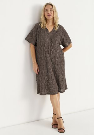 Vrouw met schouderlang blond haar, gekleed in een getextureerde donkerbruine jurk tot op de knie en zwarte enkellaarsjes met bandje, staand tegen een witte muur.