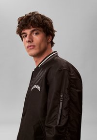 Joven con cabello rizado castaño, vistiendo una chaqueta bomber negra con cuello a rayas blancas, mirando ligeramente de lado sobre un fondo gris.