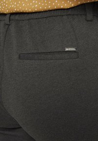 Grijze textuurbroek met een achterzak die voorzien is van een klein zilveren merklabel. Een gedeeltelijk zichtbare, gemusterde mosterdblouse erboven.