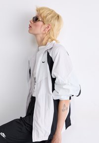Veste coupe-vent Nike en blanc et noir, dotée d'une fermeture éclair, d'une coupe décontractée, de poignets côtelés et de multiples textures. Accessoirisée avec des lunettes de soleil.