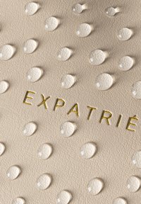 Beige nahkatuote, jossa on kohokuvioituja pisaroita ja kultainen "EXPATRIÉ" -teksti. Huomattava tekstuuri ja materiaalin yksityiskohdat parantavat kokonaisdesignia.