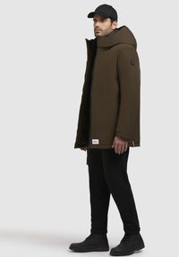 Brun isoleret parka med høj krave og hætte, sort indvendig. Sammen med sorte bukser og sorte snørebåndsstøvler.
