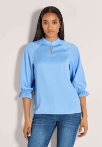 Vrouw met steil donker haar, gekleed in een lichtblauw blouse met een knoopsluiting bij de sleutelgatnek en volants op driekwartmouwen, gecombineerd met blauwe jeans.