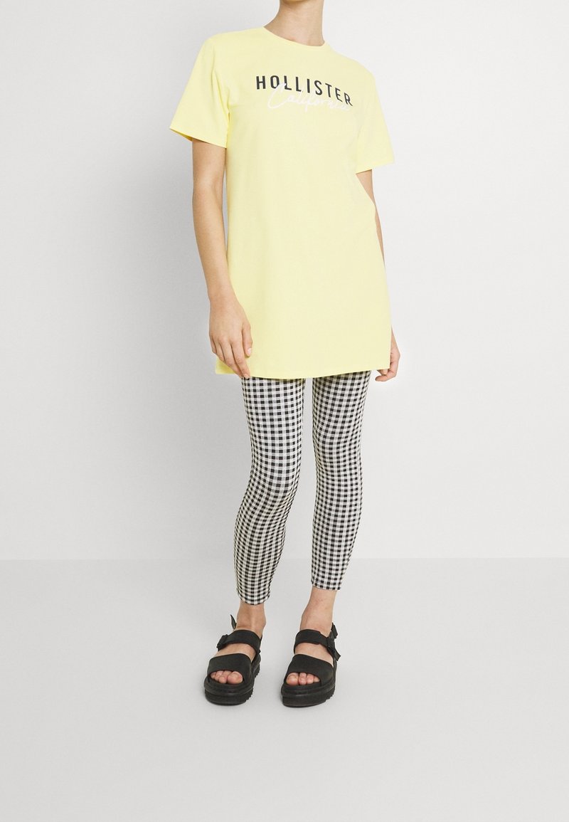 Geel katoenen t-shirt met het logo "Hollister California", gecombineerd met zwart-witte ruitleggings en zwarte plateau sandaaltjes.