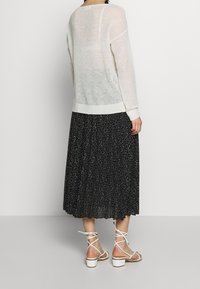 Leichter weißer Strickpullover mit gerippten Bündchen, kombiniert mit einem schwarzen, gepunkteten Faltenrock. Weiße, verzierte Sandaletten mit Absatz runden das Outfit ab.