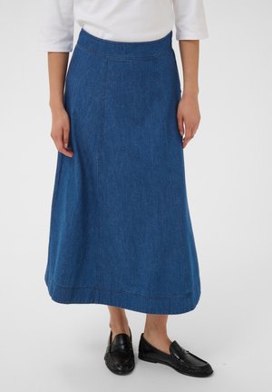 Personne portant une longue jupe en jean bleue, un haut blanc à manches trois-quarts et des mocassins noirs, debout devant un fond uni.