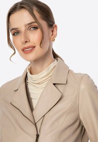 Beige leren jack met een kraag, rits aan de voorkant en subtiele stikdetails. Gecombineerd met een crème turtleneck en gouden studs.