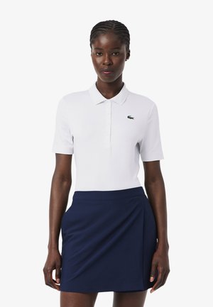 Vrouw draagt een wit poloshirt met korte mouwen en een klein logo, en een marineblauwe rok tot op de knie, staand tegen een effen achtergrond.
