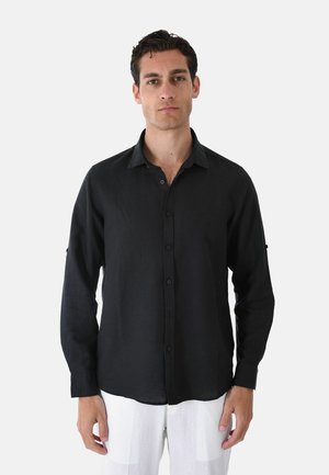 Uomo che indossa una camicia nera a maniche lunghe con bottoni e pantaloni bianchi, in piedi davanti a uno sfondo bianco uniforme.